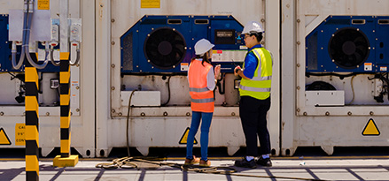 Two workers in high visibility jackets and hard hats stand by an outdoor refrigeration compressor talking.