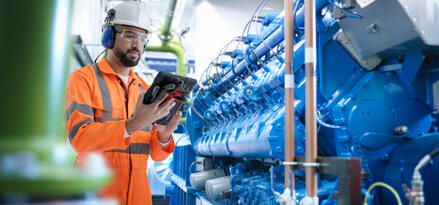 A worker in an orange high-visibility suit and hard hat is inspecting equipment in an industrial facility.