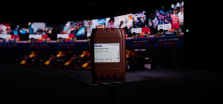 A brown 20L Mobil oil jerrycan on a small black pedestal with a blurred racetrack and screens in the background.