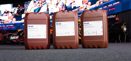 Three brown jerrycans with Mobil labels lined up on a carpeted floor in front of a large curved LED screen showing racing images