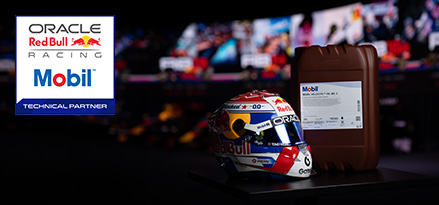 A racing helmet on display beside a large Mobil-branded oil container, with Oracle Red Bull Racing and Mobil logos visible against a blurred racetrack backdrop.