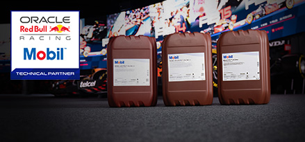 Three brown 20L Mobil oil containers lined up in front of a Red Bull Racing garage backdrop; Mobil and Oracle Red Bull Racing logos at left.