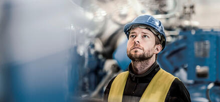 Man in a hard hat and yellow safety vest looking upward inside an industrial facility