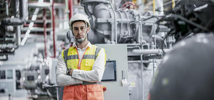 Factory worker in a hard hat and high-visibility vest standing with arms crossed in an industrial plant