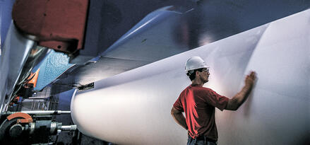 Factory worker in a hard hat inspects a large roll of paper inside industrial printing machinery.